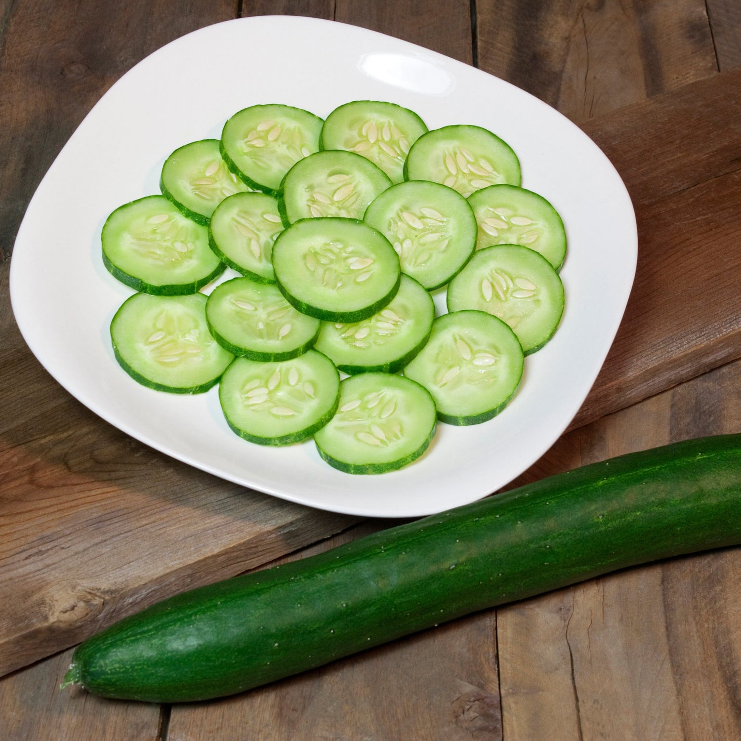 Sliced cucumbers on a white plate with a whole cucumber beside it on a wooden surface